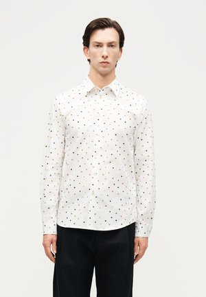 Mężczyzna stojący przodem, ubrany w białą koszulę na guziki z kolorowym, drobnym wzorem w kwadraty oraz czarne spodnie.