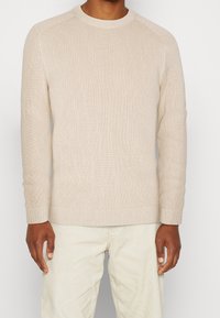 Beige ribbad tröja med långa ärmar, rundad hals, texturerad tyg och åtsittande manschetter, i kombination med ljusa byxor.