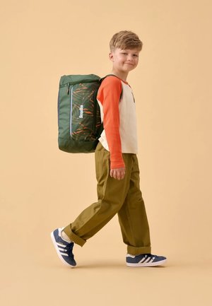 Green backpack with orange patterns, zippered compartments, adjustable straps, worn by a child in a long-sleeve white and red shirt, khaki pants, and sneakers.
