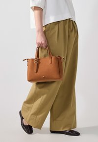 Borsa tote in pelle color cuoio con due manici corti, chiusura con zip e accenti hardware discreti; indossata con pantaloni larghi color oliva e ballerine nere.