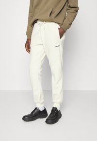 Calças de treino felpudas de cor creme com cintura elástica e bolsos laterais, apresentando um pequeno logótipo em texto preto no lado esquerdo. Combinadas com sapatos pretos.