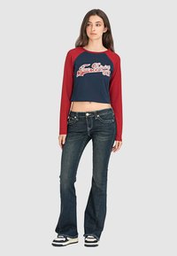 Crop-Top mit roten langen Ärmeln und navyblauem Körper, mit weißem Schriftzug. Kombination mit dunkelblauen Schlagjeans und schwarz-weißen Sneakers.