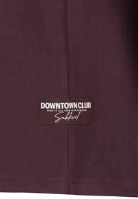 Maroon t-shirt with a smooth texture. Features a rectangular label at the bottom reading "DOWNTOWN CLUB" in white text on a dark background.