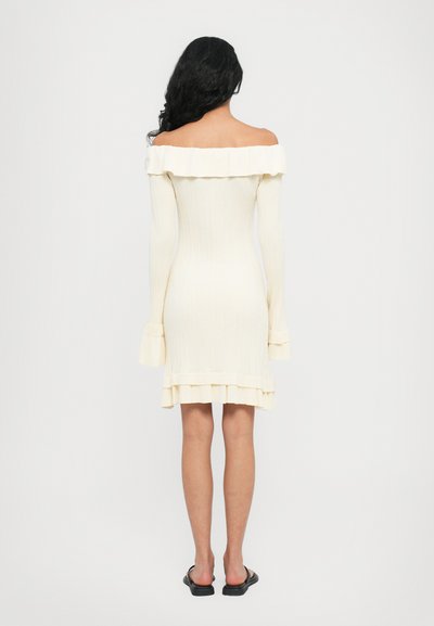 Off-the-shoulder, ribbed knit dress in cream with a ruffled neckline and hem, featuring long sleeves and a fitted silhouette.