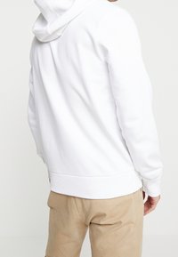 Sudadera blanca con capucha de textura suave, puños y dobladillo acanalados. Detalles de costura visibles, combinada con pantalones beige. Diseño minimalista.