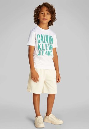 Camiseta blanca con estampado turquesa que dice "CALVIN KLEIN JEANS", combinada con pantalones cortos beige y zapatillas beige. Ajuste casual y relajado.