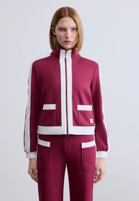 Μπορντό μπουφάν με λευκές λεπτομέρειες, κλείσιμο με φερμουάρ, όρθιο γιακά και εμπρόσθιες τσέπες. Μαλακό ύφασμα με εφαρμοστή σχεδίαση, συνδυασμένο με παντελόνι σε ίδιο χρώμα.