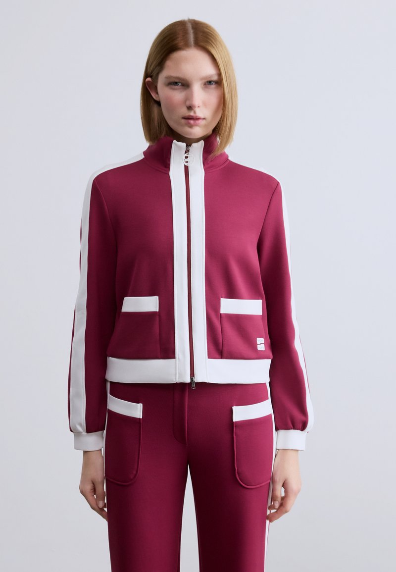 Μπορντό μπουφάν με λευκές λεπτομέρειες, κλείσιμο με φερμουάρ, όρθιο γιακά και εμπρόσθιες τσέπες. Μαλακό ύφασμα με εφαρμοστή σχεδίαση, συνδυασμένο με παντελόνι σε ίδιο χρώμα.