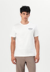 Witte katoenen t-shirt met ronde hals en korte mouwen, met een klein zwart logo aan de linkerkant. Gemaakt met een paar brown trousers.