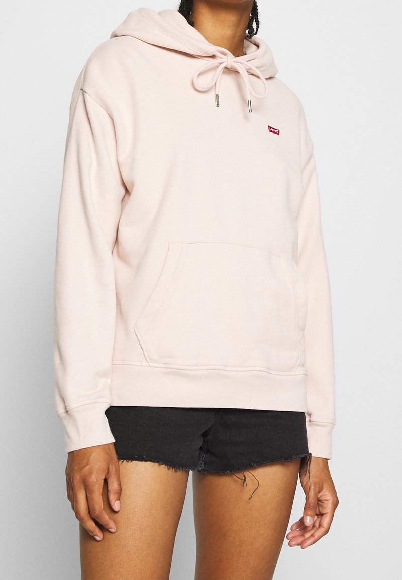 Person som bär en ljusrosa hoodie med snoddar och en framficka, ihop med svarta slitna denimshorts.