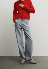 Kvinna stående med handen i fickan på ljusblå jeans, iförd röd knäppt cardigan och beige skor framför en enfärgad vit vägg.