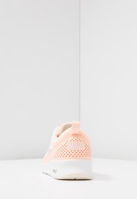 Ljus persiko Nike-sneaker sedd från baksidan, med perforerad häl och vit sula på en vit yta med mjuk grå bakgrund.