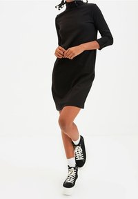 Robe noire avec un col haut et des manches trois-quarts. Tissu lisse, forme légèrement trapèze, associée à des baskets plateformes noires et des chaussettes blanches.