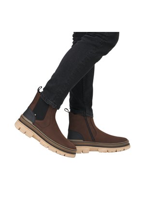Bottines en cuir marron avec des panneaux latéraux élastiques et un accent noir texturé. Elles présentent une semelle en caoutchouc blanc épais et une languette de traction.