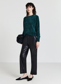 Türkiser Strickpullover, schwarze Leder-Hosen mit geradem Bein und schwarze spitze Ballerinas. Das Model hält eine schwarze Clutch. Minimalistisches Design mit strukturiertem Finish.