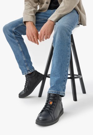 Fekete bőrből készült, magas szárú sneakers, textúrált elemekkel és narancssárga csíkkal. Kék farmernadrággal viselve, egy fekete fa széken ülve.