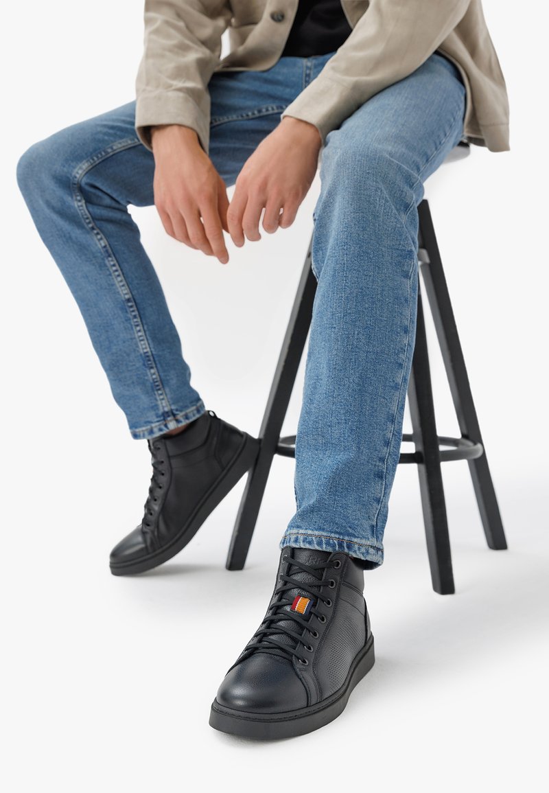 Sneakers alte in pelle nera con dettagli testurizzati e striscia arancione. Indossate con jeans blu, seduti su uno sgabello di legno nero.