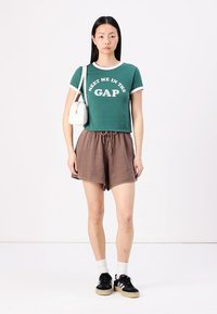 Camiseta verde corta con detalles y texto en blanco, pantalones cortos marrones texturizados con cintura elástica, bolsa blanca, zapatillas negras y calcetines blancos.