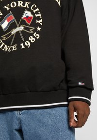 Felpa nera con logo e bandiere, con dettagli bianchi e rossi. Polsini e fondo a coste con ulteriore dettaglio di branding sulla manica.