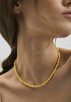 Hals und Schulter einer Frau mit einer goldenen Perlenkette, einem lose fallenden Haarstrang und einem neutralen, hellen Hintergrund.