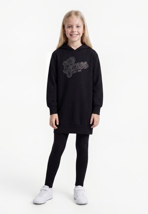 Jeune fille souriante portant une robe à capuche noire à manches longues avec l'inscription "Guess EST. 1981", des leggings noirs et des baskets blanches, debout sur fond blanc.
