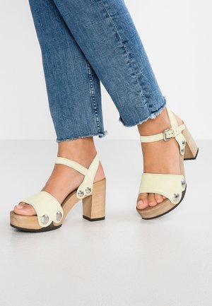 Fødder iført lysebeige plateau-sandaler med træblok hæle og metalnitter, kombineret med blå jeans med opsmøgede kanter.