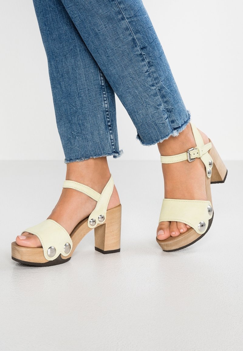 Pieds portant des sandales compensées beige clair avec talons carrés en bois et clous métalliques, associées à un jean bleu à ourlet effiloché.