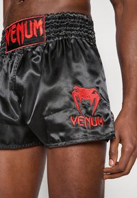 Pantalones cortos de satén negro con una cintura fruncida. Presentan el texto "VENUM" y el logo bordados en rojo. Textura suave y corte atlético.
