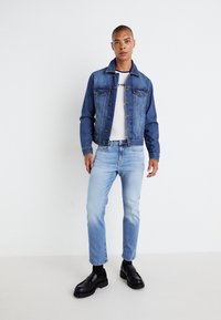 Denimjakke i mellomblå med knappelukking, kombinert med en hvit t-skjorte og lyseblå jeans. Sorte sko komplementerer antrekket.
