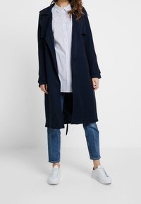 Marineblauer Trenchcoat mit tailliertem Gürtel, offenem Vorderteil und Kragen, getragen über einem leicht gestreiften Hemd, kombiniert mit Jeans und weißen Turnschuhen.