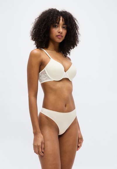 Γυναίκα με κατσαρά μαλλιά που φοράει λευκό δαντελωτό σουτιέν και αντίστοιχο λευκό εσώρουχο, στέκεται μπροστά σε απλό φωτεινό φόντο.