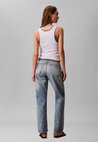 Γυναίκα που στέκεται με την πλάτη προς τα εμπρός, φορώντας λευκό τοπ με λεπτή στρώση πάνω από γκρι μπλούζα, ξεβαμμένο μπλε τζιν με διακριτικό μοτίβο και μαύρα σανδάλια.