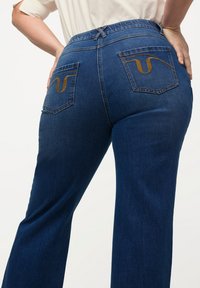Los jeans de denim azul oscuro tienen una cintura alta, piernas anchas y dos bolsillos traseros con un distintivo bordado amarillo en un patrón de ola.