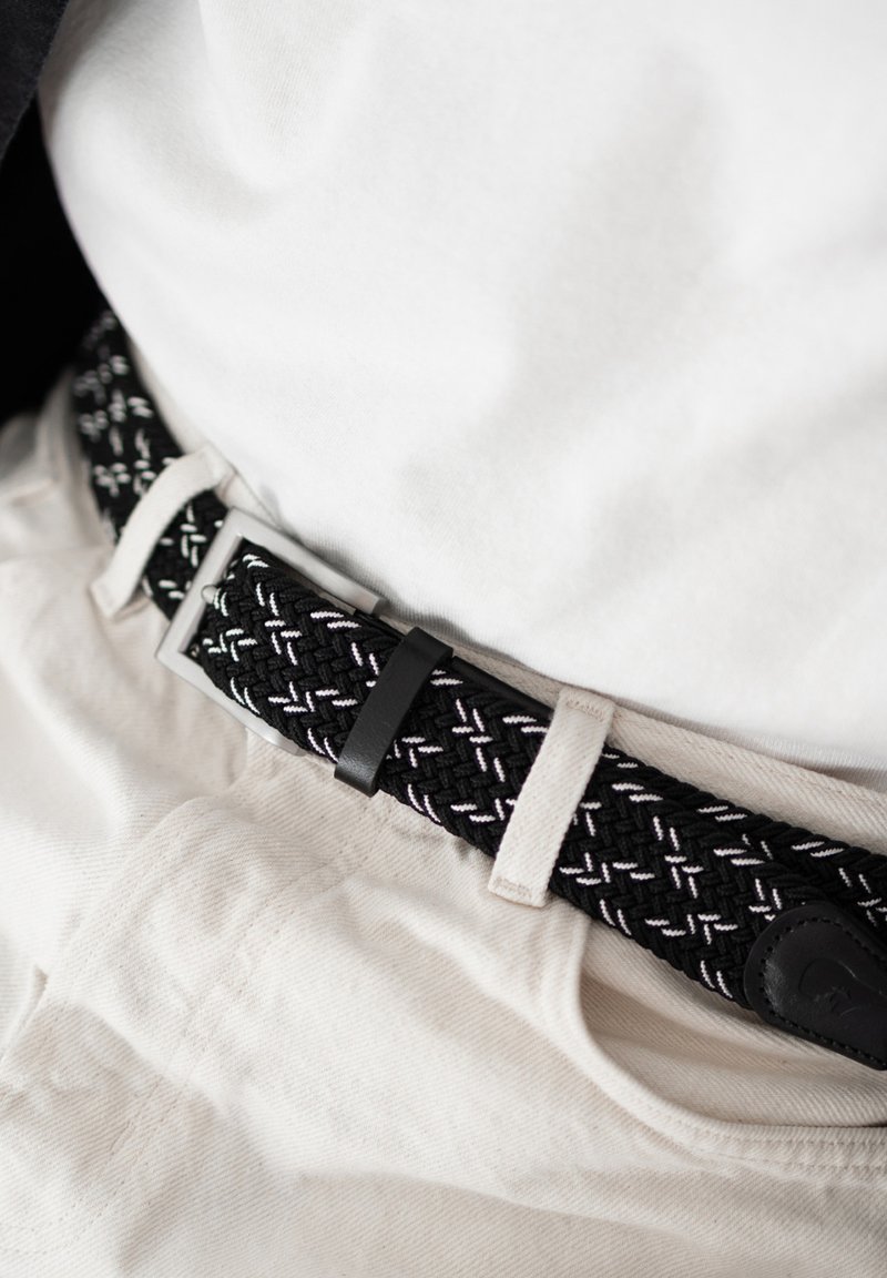 Ceinture tissée noire et blanche attachée autour d'un pantalon beige porté avec une chemise blanche rentrée.