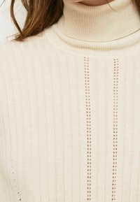 Pull à col roulé crème présentant une texture côtelée verticale avec de petits accents en openwork le long du centre, en tissu tricoté doux.