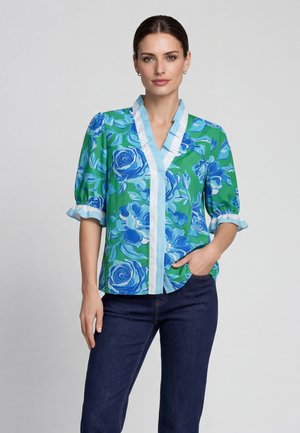 Donna con capelli scuri che indossa una blusa verde con stampa floreale blu e bordi azzurri, jeans blu scuro, in piedi con la mano in tasca.