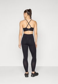 Svart sport-bh med kryssryggdesign og logo, kombinert med høyhårede svarte leggings. Modellen har på seg svarte joggesko.