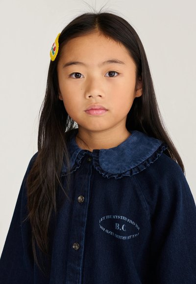 Jeune fille aux longs cheveux foncés portant une chemise bleu foncé boutonnée avec un col à volants et une barrette colorée sur le côté gauche.