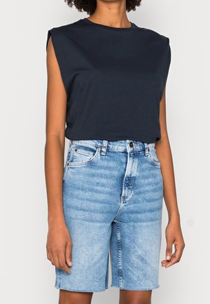 Person iført sort ærmeløs top og lyseblå denimshorts, stående mod en ensfarvet lys baggrund.