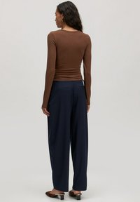 Vrouw in een bruine long-sleeve top, marineblauwe wijdpijpen broek en bruine hakken, staande met haar rug naar de camera tegen een lichte, effen achtergrond.