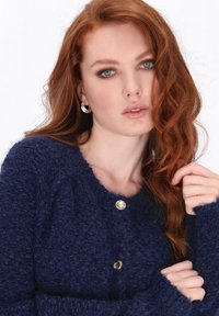 Red-haired woman with wavy hair wears silver hoop earrings and a textured navy blue sweater with gold buttons, holding a strand of hair.