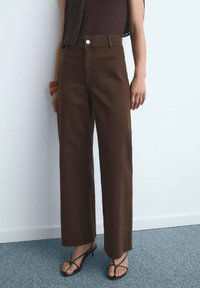 Pantalones anchos marrones hechos de una tela texturizada, con una cintura alta, bolsillos frontales y un dobladillo ligeramente deshilachado. Combinados con sandalias negras de tiras.