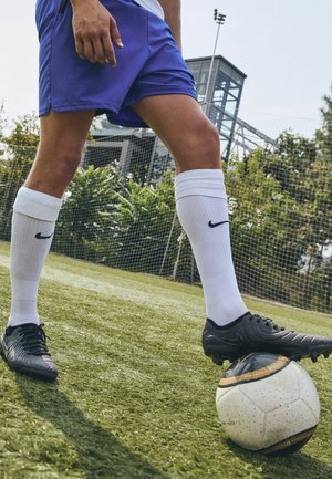 Joueur de football portant un short bleu, des chaussettes blanches et des crampons noirs contrôle un ballon de football blanc et or sur un terrain en herbe.