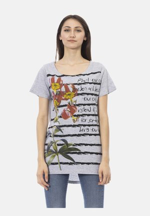 Femme portant un t-shirt gris à manches courtes avec des rayures noires, un imprimé floral rouge et jaune, et un texte manuscrit partiellement lisible, associé à un jean bleu.