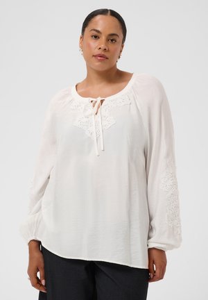 Femme aux cheveux foncés attachés en arrière, portant un chemisier blanc ample avec des détails en dentelle sur la poitrine et les manches, ainsi qu'un pantalon noir.