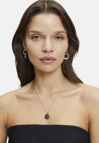 Zilveren ketting met een druppelvormige zwarte edelsteen hanger. Eenvoudig ontwerp van de ketting. Model draagt zilveren ringoorbellen en een strapless top.