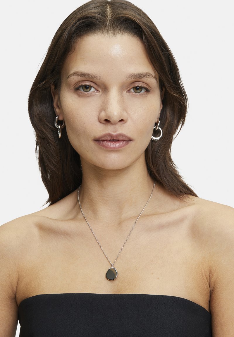 Zilveren ketting met een druppelvormige zwarte edelsteen hanger. Eenvoudig ontwerp van de ketting. Model draagt zilveren ringoorbellen en een strapless top.