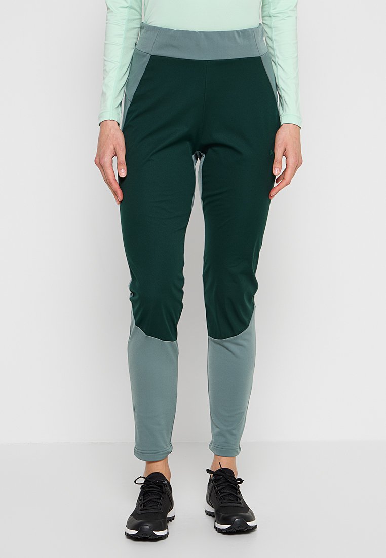 Personne portant un pantalon de sport slim bicolore vert foncé et gris avec des chaussures de course noires, se tenant devant un fond blanc.