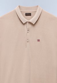 Polo di colore beige con colletto, tre bottoni e una piccola bandiera della Norvegia ricamata sul lato sinistro del petto. Tessuto morbido, design minimal.