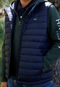 Granatowa pikowana kamizelka z zamkiem błyskawicznym i bocznymi kieszeniami, noszona na zielonej bluzie z kapturem. Na lewej piersi znajduje się logo, a materiał ma gładką teksturę.